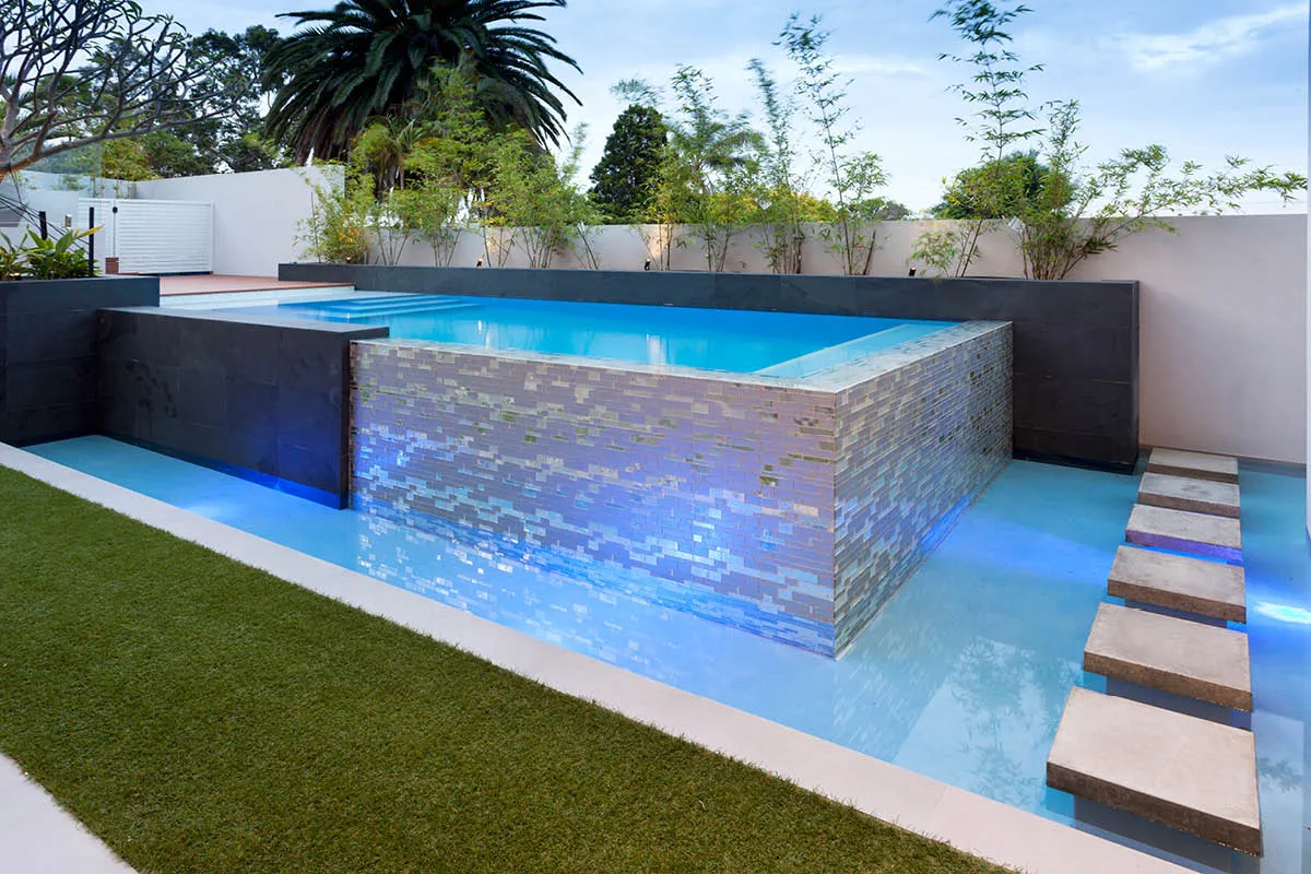 Raised stone clad pool with stepping pavers across artificial lawn