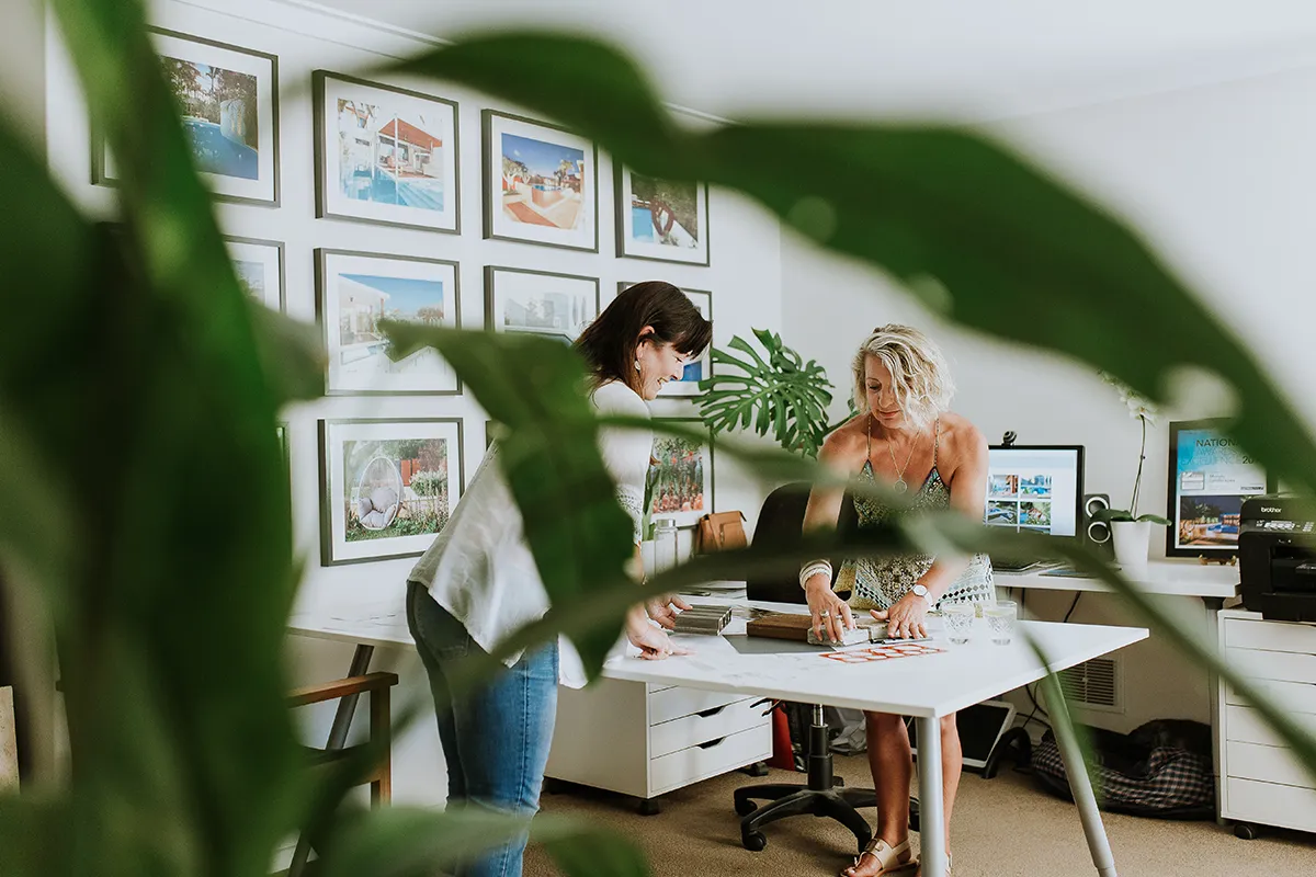 Two women working at a desk in a bright office with framed prints behind