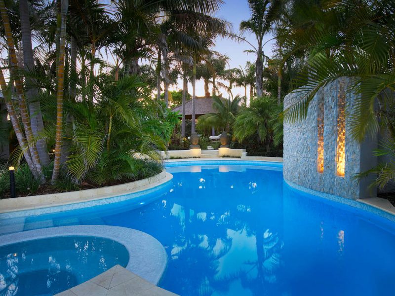 Turquoise blue kidney shape pool surrounded by lush tropical garden with palms that pavilion in rear with large stone busts and stone water feature with light channels the pool is tiled in white mosaic