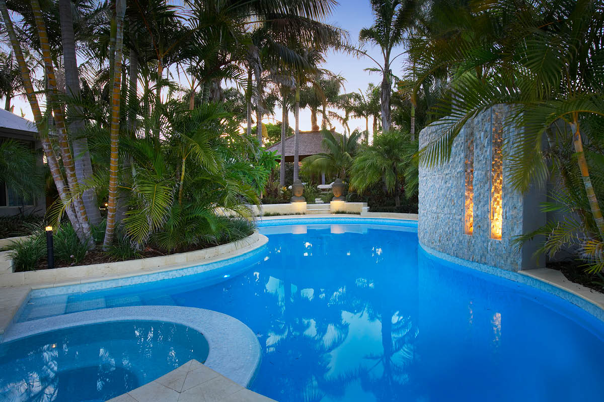 Turquoise blue kidney shape pool surrounded by lush tropical garden with palms that pavilion in rear with large stone busts and stone water feature with light channels the pool is tiled in white mosaic