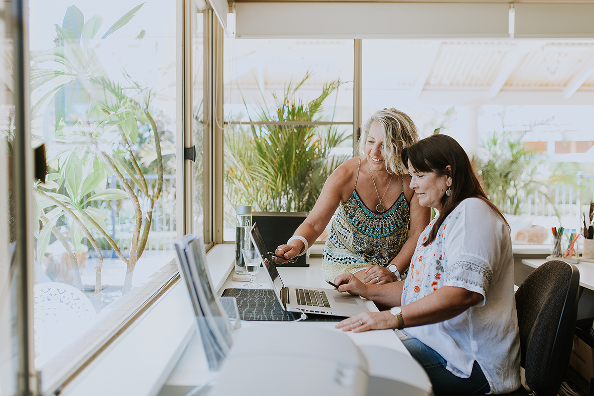 Blonde woman pointing at laptop computer being operated by dark haired women in home office