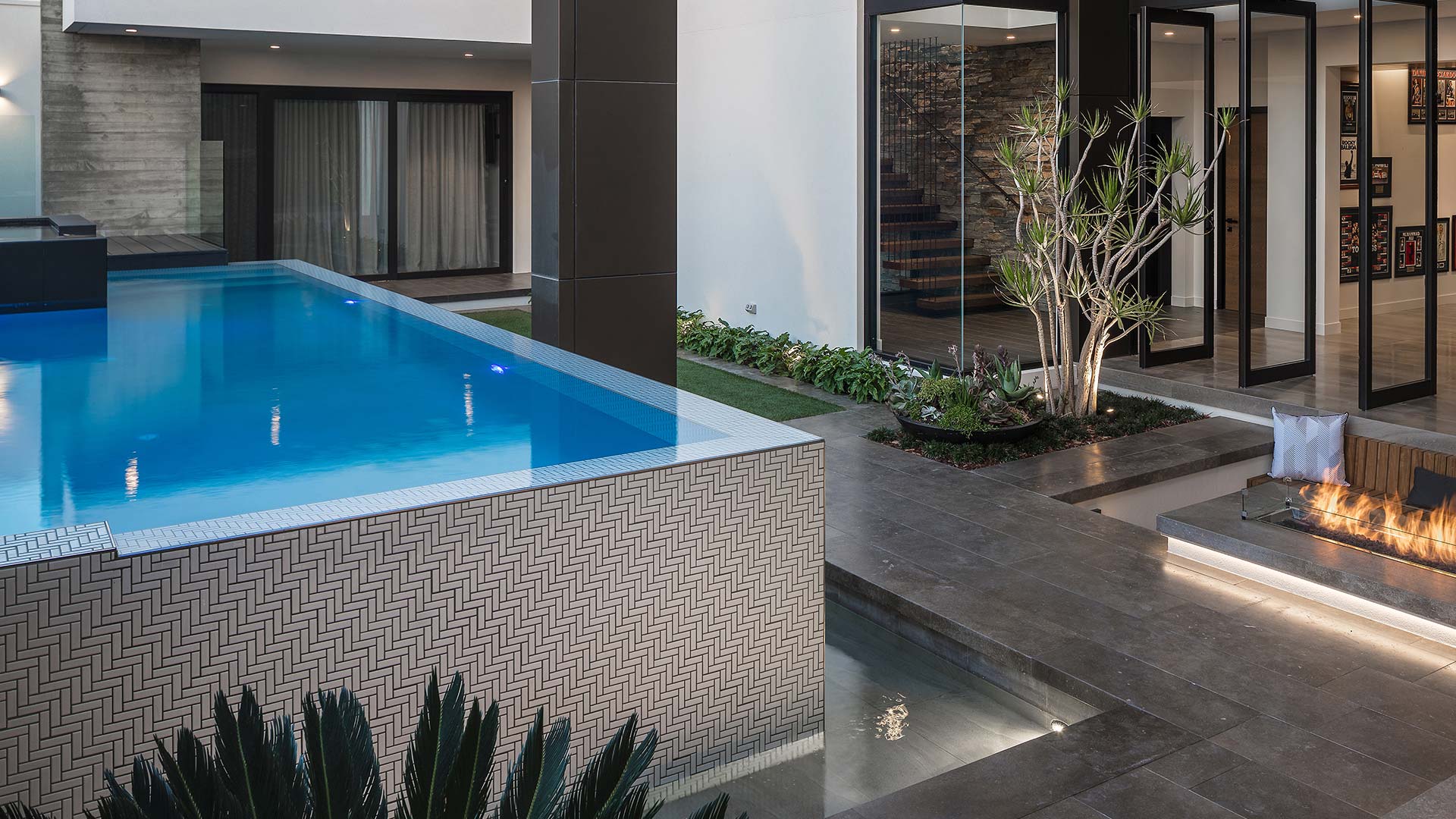 Custom designed infinity edge swimming pool, with herringbone mosaic water edge. Custom design gas fire pit with timber seat. Natural stone paving and wall enhanced with soft planting.